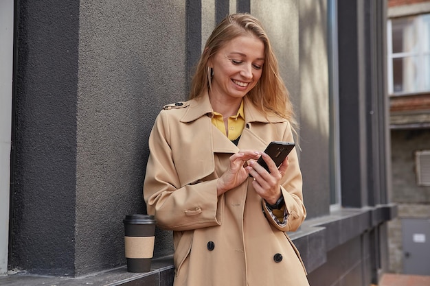 Caucasian woman in beige trench coat using smartphone outdoors on sunny day