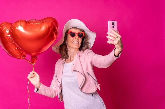 A caucasian woman having fun with a white hat in a nightclub with some heart balloons taking a selfie with her mobile phone pink background