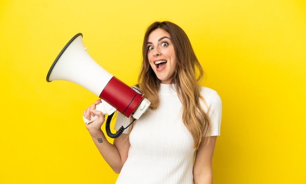 Caucasian woman isolated on yellow background holding a megaphone and with surprise expression