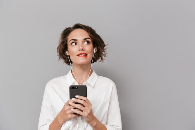 caucasian woman listening to music on smartphone via earphones, isolated over gray wall