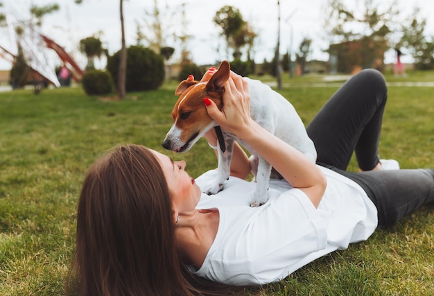 Caucasian woman plays with her dog lying on the grass in the park The concept of animals friendship people and love a woman lies on the grass and plays with a jack russell terrier