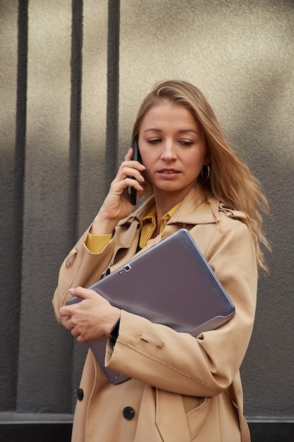 caucasian woman in stylish beige coat calling by smartphone holding laptop