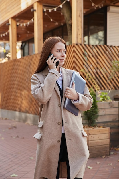 Caucasian woman in stylish beige coat calling by smartphone holding laptop