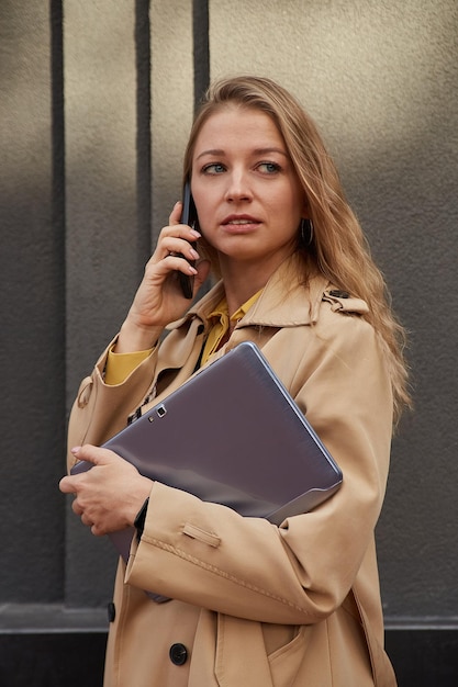 Caucasian woman in stylish beige coat calling by smartphone holding laptop