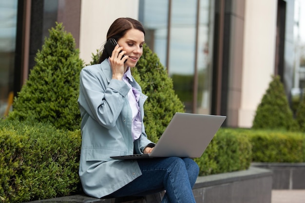 Caucasian woman using laptop and talking on the phone in the city outdoors Distant work Modern business woman Uses gadgets Lifestyle