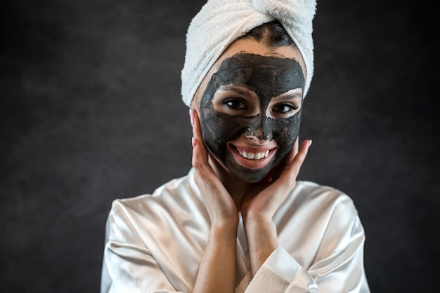 Caucasian woman wear white towel with black clay mud nourishing mask on face isolated on black