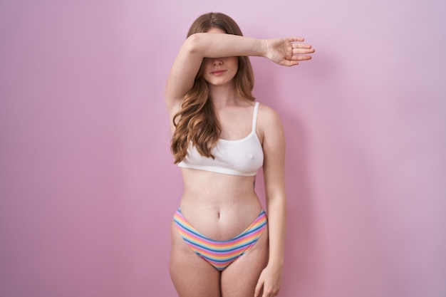 Caucasian woman wearing lingerie over pink background covering eyes with arm, looking serious and sad. sightless, hiding and rejection concept