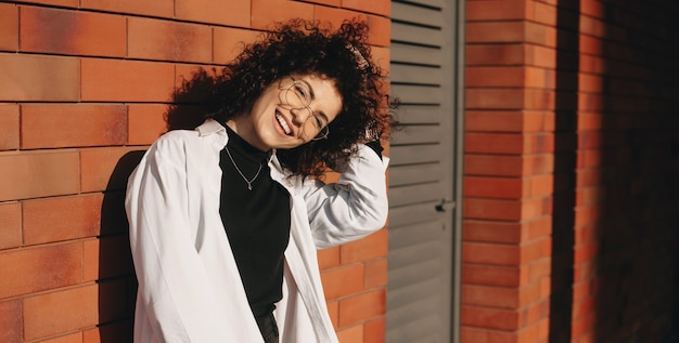 Caucasian woman with curly hair posing on a orange stone wall with eyeglasses smiling at camera