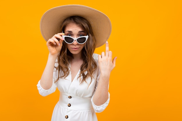 Caucasian woman with hat and sunglasses is posing on camera on yellow