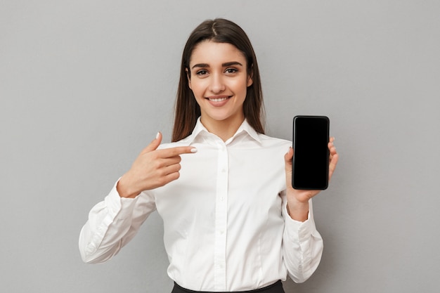 Caucasian woman with long brown hair doing commercial and presenting black copyspace screen of mobile phone , isolated over gray wall