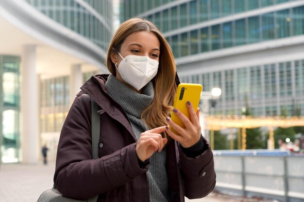 Caucasian woman with winter clothes is typing with finger chatting with someone on smartphone while wearing a protective mask outside