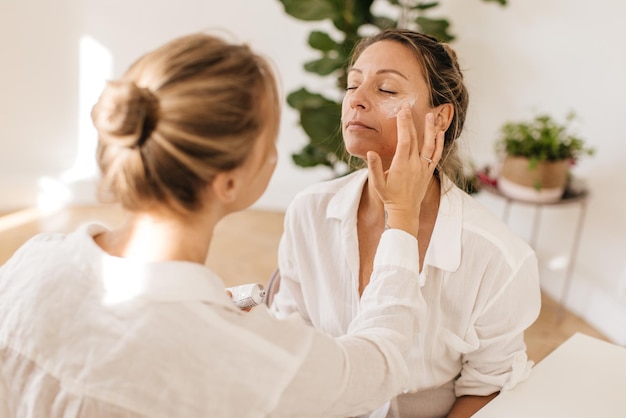 Caucasian young blonde girl applies moisturizer on her mother's face while sitting side by side in light room. Wellness and self care concept