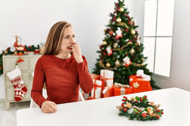 Caucasian young blonde woman sitting on the table by christmas tree looking stressed and nervous with hands on mouth biting nails. anxiety problem.