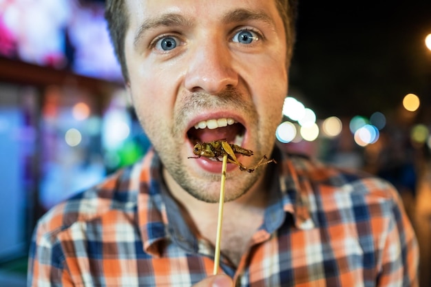 Caucasian young male eating cricket at night market in Thailand