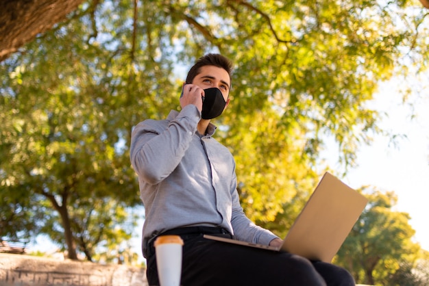 Caucasian young man wearing a face mask working and studying in engineering talking on the phone and using his laptop in a beautiful park on a sunny day..