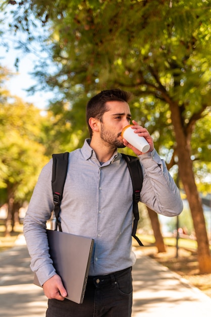 Caucasian young student and worker man drinking a takeaway coffee and carrying his laptop in a beautiful park on a sunny day.. Vertical