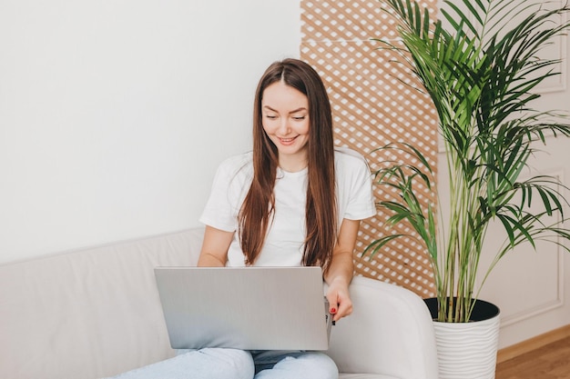 Caucasian young woman looking at laptop screen sitting on sofa working and smiling