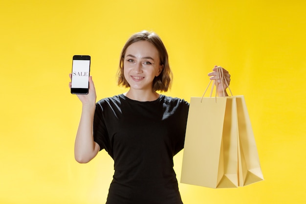 Caucasian young woman smiling and holding mobile phone, and shopping package and looking into phone screen