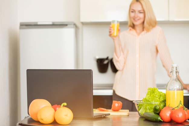 Caucasian young woman wearing striped shirt holding glass of fresh juice standing in kitchen behind the table with laptop computer, vegetables and fruits, watching the show while cooking.