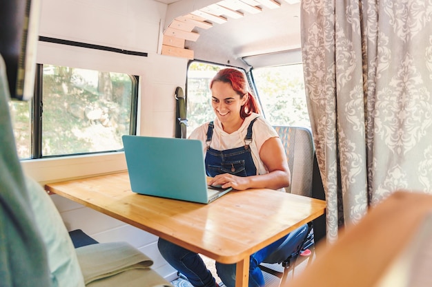 Caucasian young woman working on her laptop in her handmade camper van while traveling Van road trip holiday and outdoor summer adventure Nomad lifestyle concept