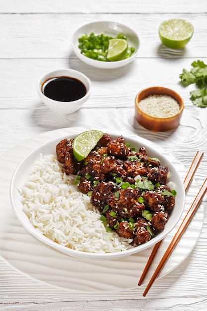 Cauliflower wings with teriyaki sauce - vegan crunchy baked cauliflower florets coated in brown sauce topped with fresh cilantro, sesame seeds, served with lime and rice on a while bowl, top view