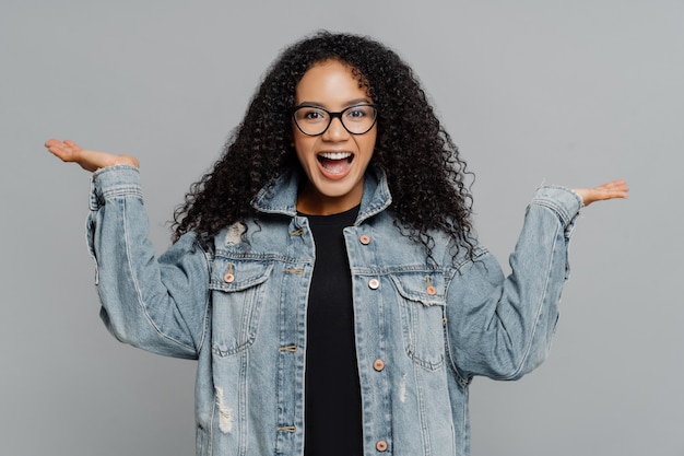 Ccurly African American woman raises palms, pretends holding something, wears optical glasses and denim jacket