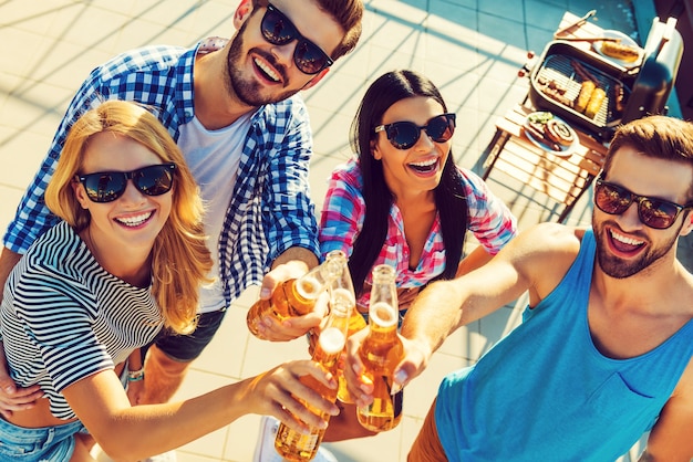 Celebrating their friendship. Top view of four cheerful young people clinking glasses with beer and looking at camera while standing outdoors