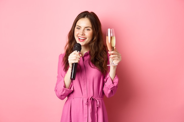 Celebration and holidays concept. Beautiful young woman saying toast, raising glass of chamapgne while making speech, holding microphone, standing over pink wall.