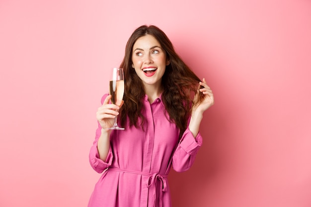 Celebration and holidays concept. Stylish coquettish woman in trendy dress, holding glass of champagne at party, smiling pleased, standing over pink wall. Copy space