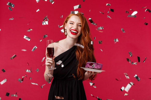 Celebration, holidays and party concept. Portrait of carefree beautiful young redhead woman having fun at birthday, celebrating laughing while confetti flying around, hold glass champagne and cake