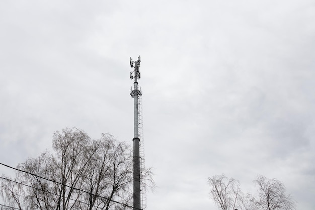 Cell tower on background of gray sky electric wires and tree branches