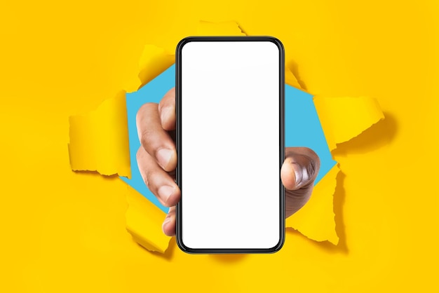 Cellular app Male hand showing smartphone with white blank screen breaking through torn yellow paper background mockup