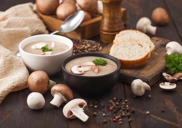 Ceramic bowl plates of creamy chestnut champignon mushroom soup with spoon, pepper and kitchen cloth on dark wooden board.