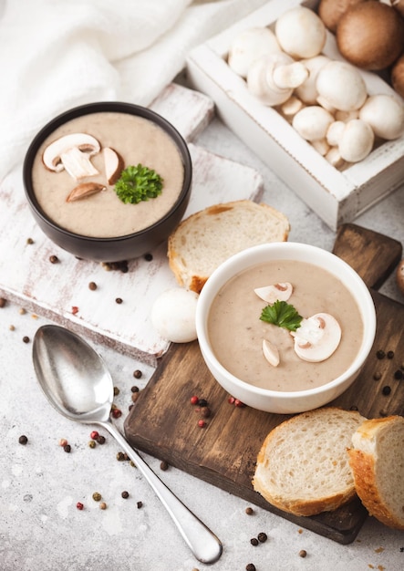 Ceramic bowl plates of creamy chestnut champignon mushroom soup with spoon pepper and kitchen cloth on white kitchen background and box of raw mushrooms Top view