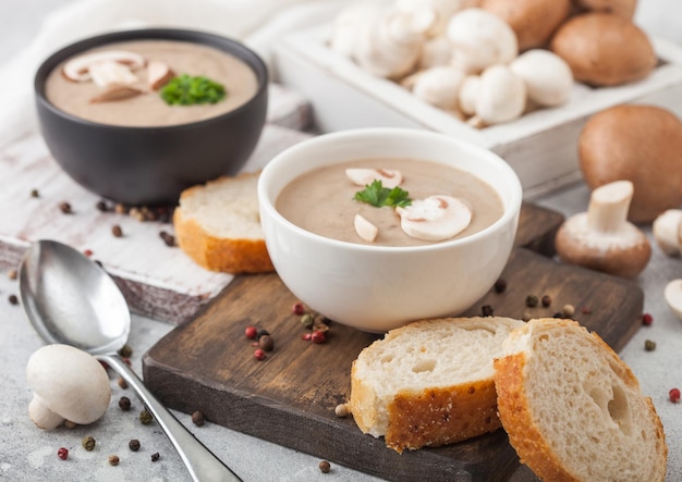 Ceramic bowl plates of creamy chestnut champignon mushroom soup with spoon pepper and kitchen cloth on white kitchen background and box of raw mushrooms