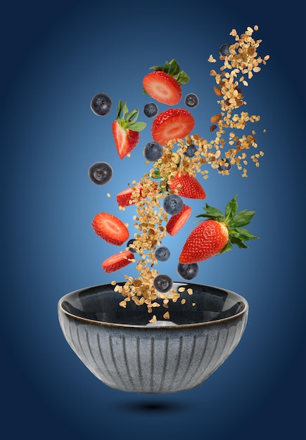 A ceramic bowl in which pieces of strawberries blueberries and muesli float in the air and fall creating a feeling of dynamics and freshness on a gradient blue background