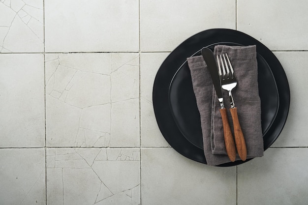 Ceramic empty black plate silverware and linen kitchen towel napkin on old ceramic tile table background Cooking stone backdrop Top view with copy space Flat lay