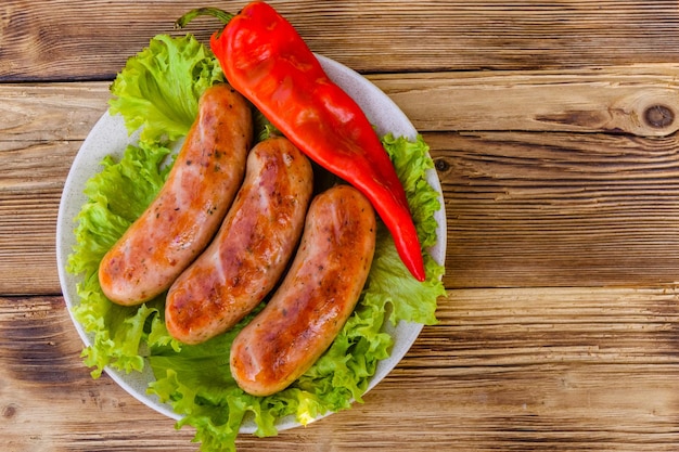 Ceramic plate with grilled sausages, pepper and lettuce leaves on rustic wooden table. Top view