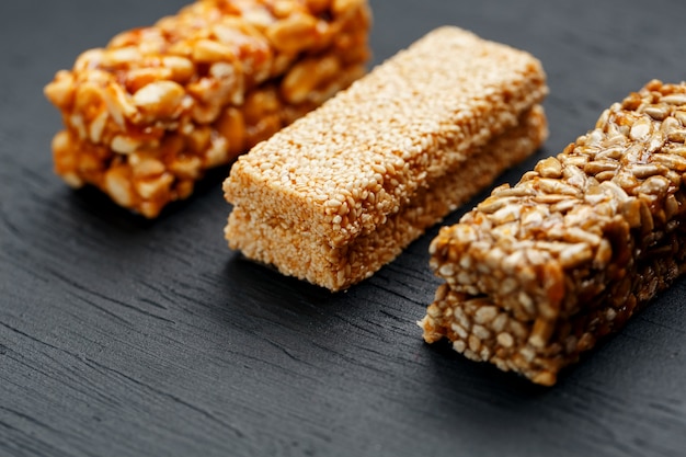 Cereal granola bar with peanuts, sesame and sunflower seeds on a cutting board on a dark stone table. View from above. Three Assorted Bars