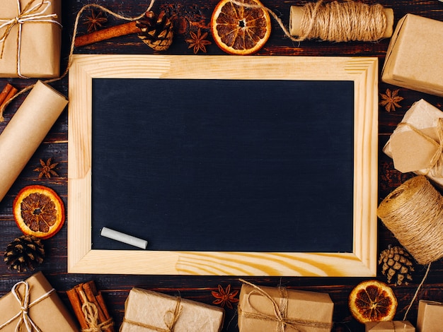 Chalk board in the middle of gifts of dried orange, cinnamon, pine cones, anise on a white table Christmas billet, save space, top view, place for text.