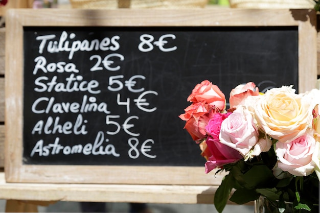 Chalkboard with the different prices of flowers written in Spanish (tulips, roses, statice, carnation, wallflower, Astromelia) and a bouquet of roses in a street flower market