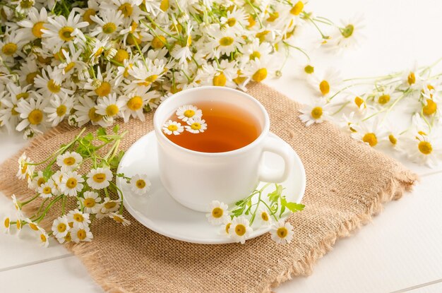 Chamomile tea in a white cup on a white wooden background with a bouquet of daisies