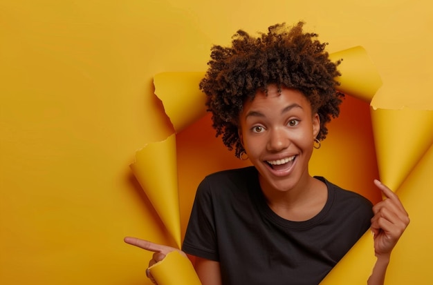 Charismatic joyful Afro woman with curly hairstyle turns away looks on right side wears fashionable shirt stands in torn paper hole