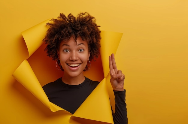 Charismatic joyful Afro woman with curly hairstyle turns away looks on right side wears fashionable shirt stands in torn paper hole