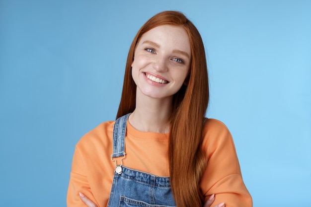 Charismatic kind pleasant redhead girl blue eyes smiling friendly listen politely customer standing blue background tilting head amused grinning cross hands chest professional confidence pose