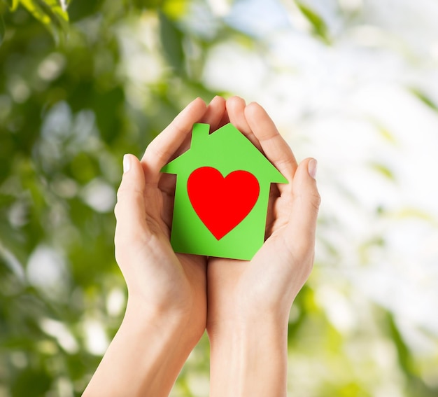 charity, real estate and family home concept - closeup picture of female hands holding green paper house with red heart