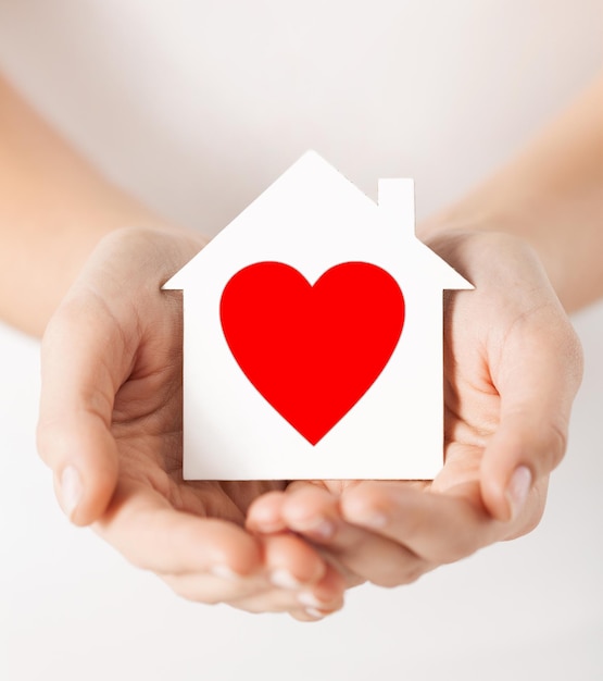 charity, real estate and family home concept - closeup picture of female hands holding white paper house with red heart