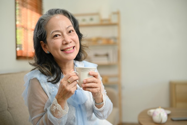 Charming 60s aged Asian woman is in the living room holding a cup of tea smiling