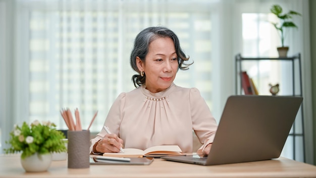 Charming 60s senior Asian female writer working at her desk writing something on her book