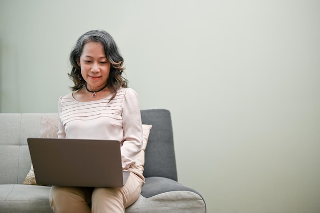 Charming agedasian woman using laptop sitting on a couch in her living room